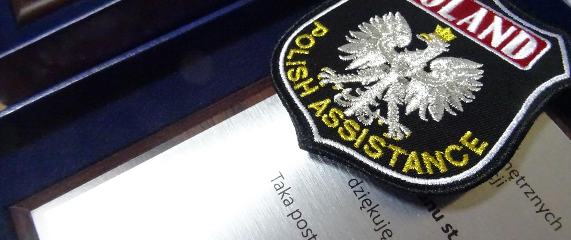 Zdjęcie przedstawia pamiątkowy ryngraf oraz tarczę strażaków, którzy wyruszają z pomocą za granicę w ramach misji.