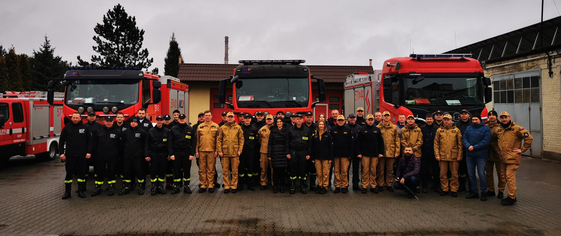 Zdjęcie przedstawia strażaków ustawionych na tle samochodów pożarniczych. Strażacy ubrani w różnego rodzaju umundurowanie