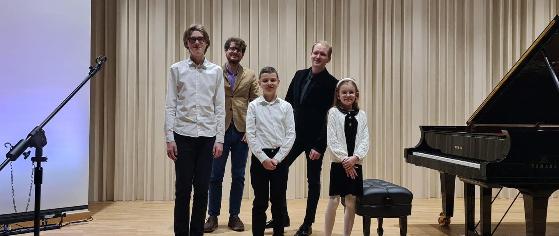 Na zdjęciu w pierwszym rzędzie trzech uczniów- dwóch chłopców i jedna dziewczynka. Chłopcy w białych koszulach i czarnych spodniach. Dziewczynka w okularach w czarnej sukience i białym sweterku. W tyle za nimi dwóch mężczyzn. Jeden w brązowej marynarce i niebieskiej koszylu, a drugi ubrany na czarno.Obok nich po prawej stronie stoi czarny fortepian.