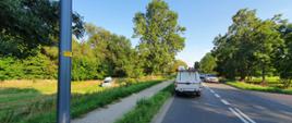 Zdjęcie ulicy, na pasie pojazd Pogotowia Energetycznego. W tle wrak pojazdu na zielonej trawie. Zdjęcie w ciągu dnia. Okres letni.