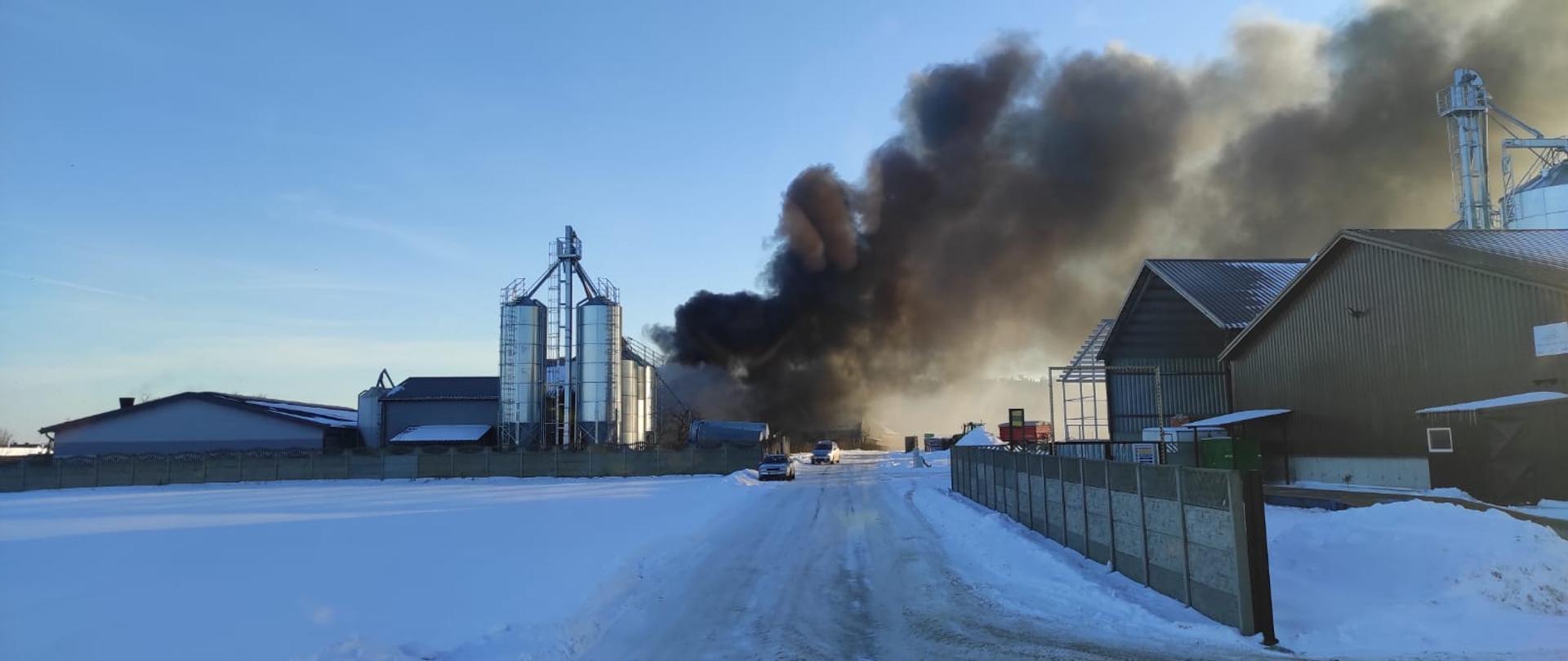 Zdjęcie zrobione w trakcie dojazdu do pożaru, widać kłęby dymy wydostające się z budynku gospodarczego z bezpośrednim sąsiedztwie z budynkami inwentarskimi. Widać duże silosy zbożowe przy budynku inwentarskim droga do pożaru utrudniona ,oblodzona powierzchnia drogi. Po drugiej stronie pożaru stoją wiaty metalowe które nie są zagrożone pożarem. 
