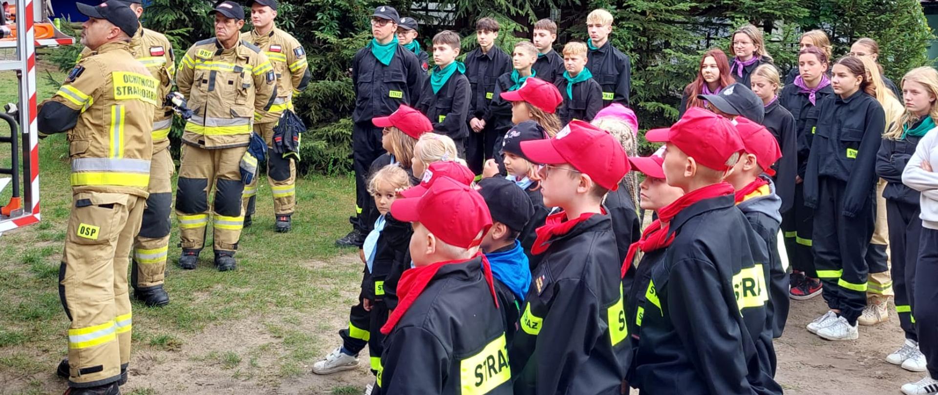 Zdjęcie przedstawia strażaków oraz dzieci i młodzież na wakacyjnym obozie strażackim.
W tle ośrodek wypoczynkowy.
