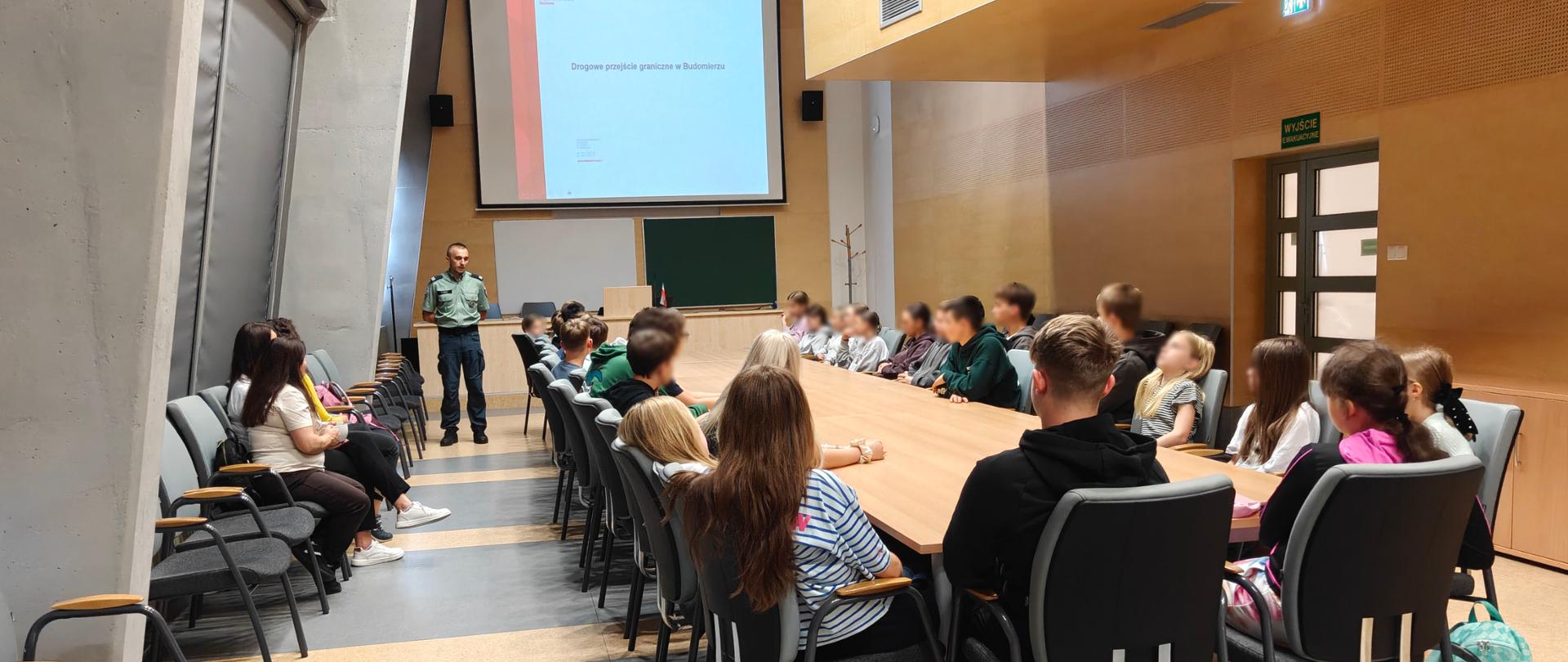 Uczniowie siedzą przy długim stole. Przed nim stoi funkcjonariusz SCS
