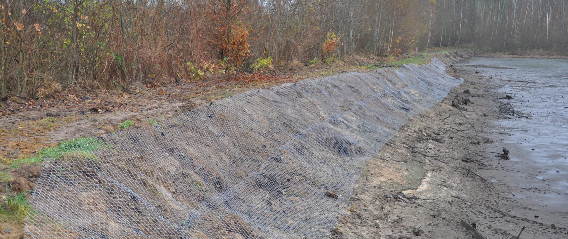 Na zdjęciu brzeg zbiornika wodnego zabezpieczony metalową siatką wbitą w zbocze drewnianymi kołkami.