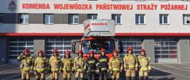 Strażacy PSP w umundurowaniu specjalnym stoją przed pojazdami pożarniczymi koloru czerwonego, które stoją przed remizami JRG PSP. Strażacy oddają hołd poległym strażakom w Ukrainie.