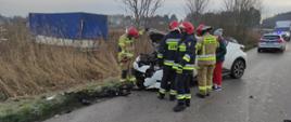 Zdjęcie przedstawia rozbity samochód po udziale w kolizji trzech samochodów. Strażacy zabezpieczają miejsce zdarzenia, w tle widać samochód ostródzkiej policji oraz samochód dostawczy czekający na przejazd.