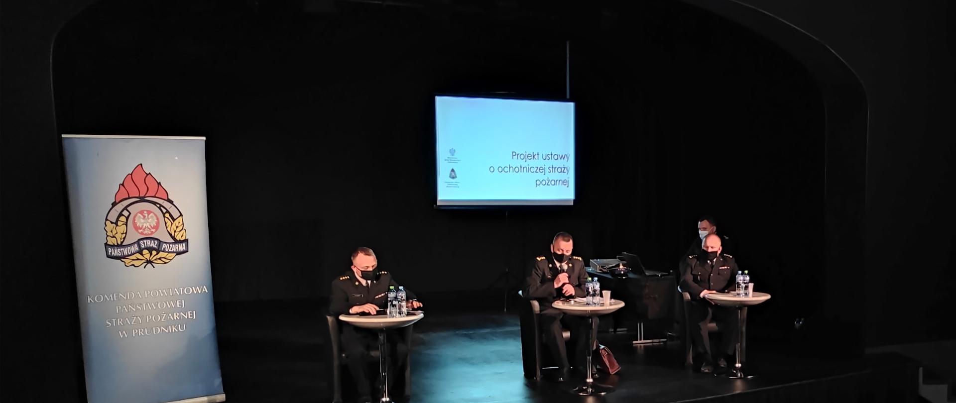 Zdjęcie przedstawia trzech funkcjonariuszy PSP w mundurach wyjściowych siedzących przy białych stolikach, po lewej stronie rollup z logiem Państwowej Straży Pożarnej, z tyłu biały ekran z widocznym tekstem Projekt ustawy o ochotniczej straży pożarnej, całość zdjęcia na czarnym tle.