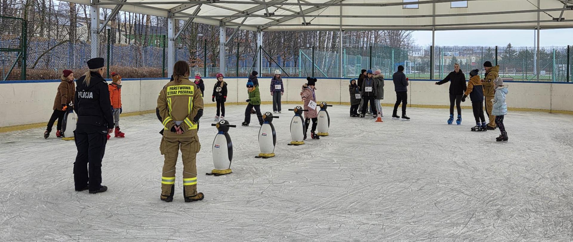 Zdjęcie przedstawia grupę osób na zadaszonym lodowisku, gdzie odbywają się zajęcia edukacyjne dotyczące bezpieczeństwa. Na pierwszym planie, tyłem do obiektywu, stoją dwie funkcjonariuszki służb mundurowych: jedna w czarnym mundurze z napisem „POLICJA” oraz druga w piaskowym stroju bojowym z napisem „PAŃSTWOWA STRAŻ POŻARNA”. Ich obecność sugeruje akcję profilaktyczną skierowaną do najmłodszych, mającą na celu naukę bezpiecznych zachowań podczas zimowego wypoczynku.
W centralnej części tafli lodu widać liczną grupę dzieci w różnokolorowych strojach zimowych. Niektóre z nich pewnie poruszają się na łyżwach, podczas gdy inne korzystają z niebiesko-białych chodzików w kształcie pingwinów, które ułatwiają naukę jazdy i pomagają utrzymać równowagę. Całość odbywa się wewnątrz dużej konstrukcji namiotowej z białym poszyciem, która chroni uczestników przed warunkami atmosferycznymi, zapewniając komfortową przestrzeń do zabawy i nauki.

