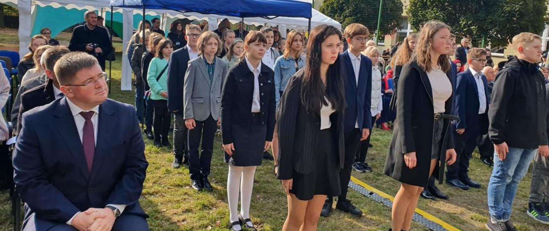 Wiceminister Tomasz Rzymkowski uczestnikiem uroczystości nadania imienia "Lotników Łaskich" szkole w Teodorach, wiceminister Tomasz Rzymkowski siedzi, obok stoją uczniowie ze szkoły w Teodorach.