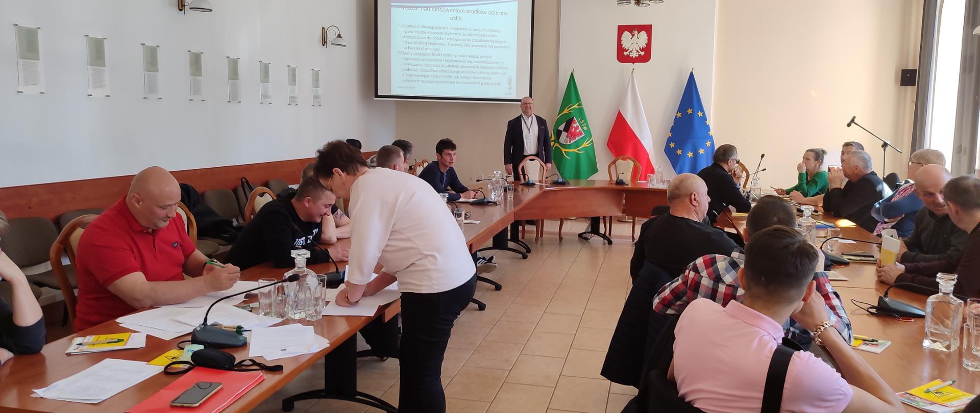 Uczestnicy szkolenia siedzą przy stole na którym znajdują się dokumenty, notatniki,
długopisy oraz dzbanki z wodą. W tle znajduje się ekran z wyświetloną prezentacją oraz
osoba prowadząca prezentację. Na ścianie widoczne są flagi i godło państwowe