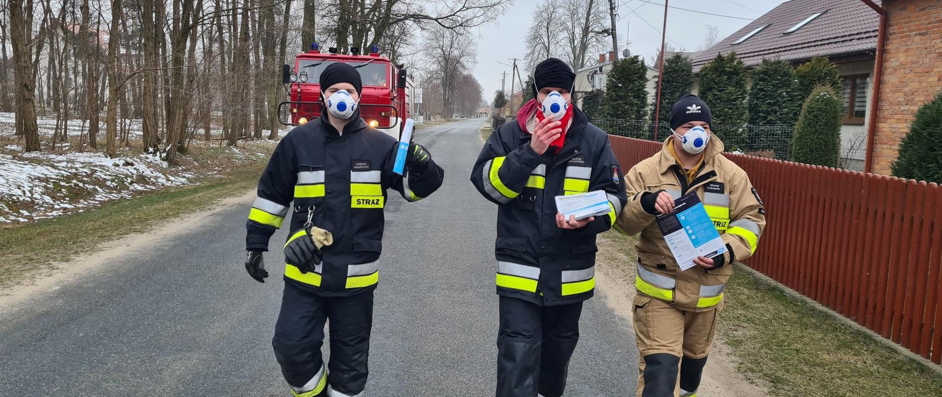 Zdjęcie przedstawia trzech strażaków ochotniczej straży pożarnej w umundurowaniu specjalnym i maseczkach na twarzy idących drogą i trzymających w ręku ulotki. Z tyłu za strażakami jedzie samochód ratowniczo - gaśniczy.