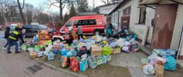 Dary przekazane przez mieszkańców Milanówka doraźnie składowane przed strażnicą OSP Milanówek przed dalszą ich segregacją w tle bus strażacki i osoby segregujące dary.