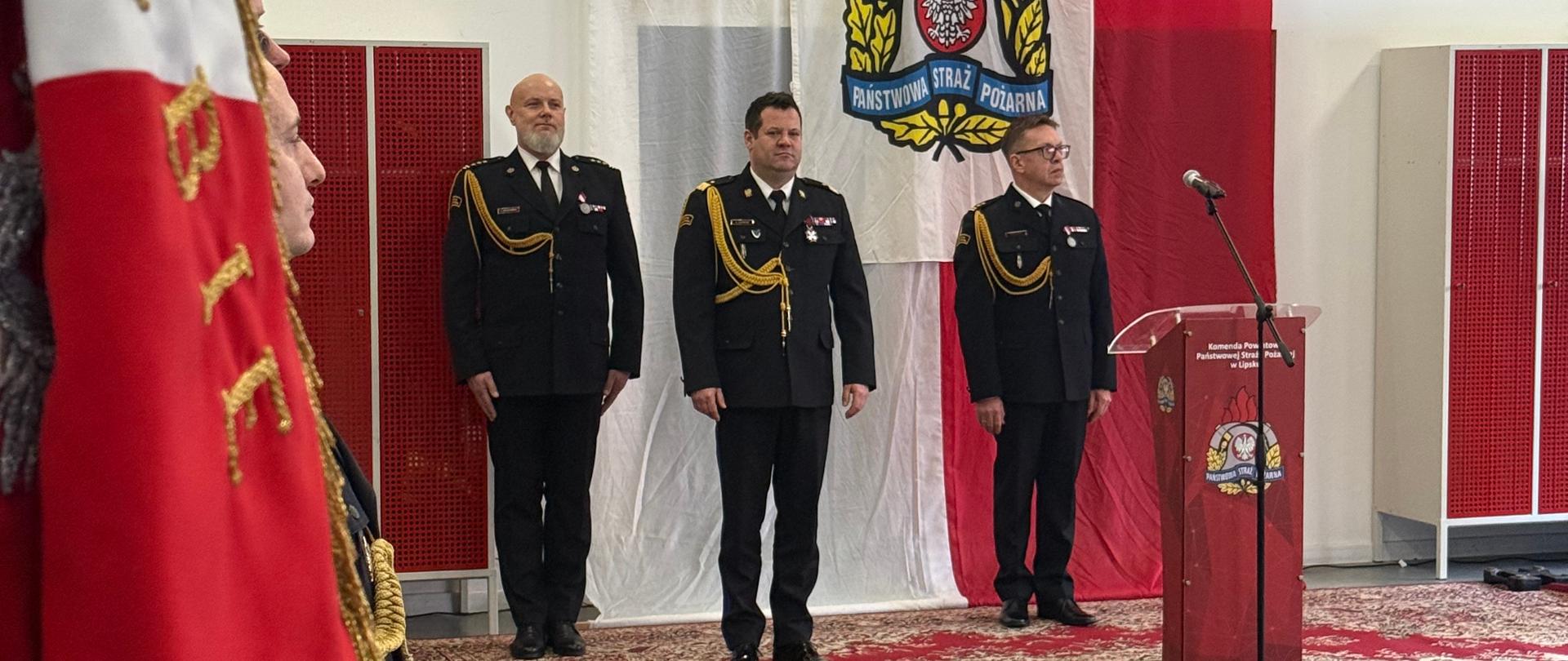 Trzech strażaków stoi przy mównicy na tle dużej biało-czerwonej flagi i logotypu PSP