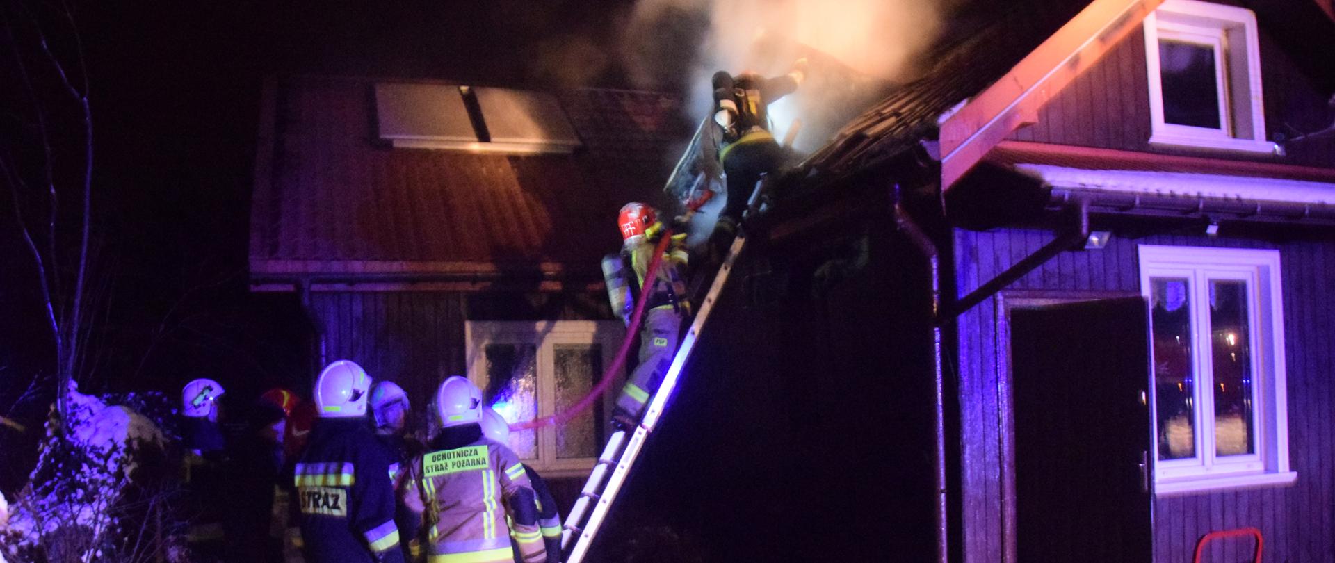 Zdjęcie przedstawia drewniany budynek mieszkalny, parterowy, strażacy wchodzą po drabinie z prądem wody