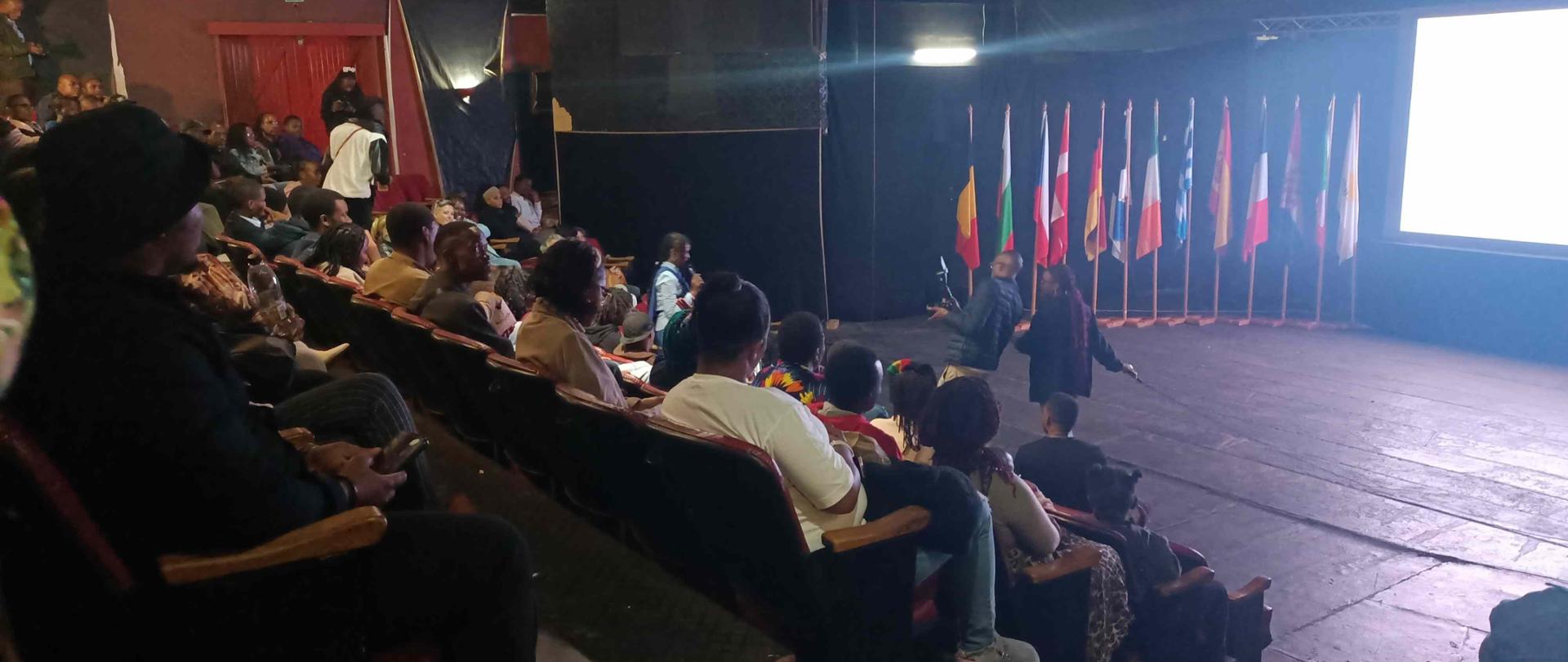 The audience at the Mbabane Theatre Club; gathered spectators in the foreground; EU flags in the background; a photographer taking photos of the audience; dim lights.