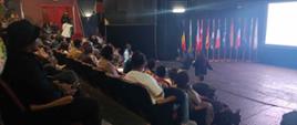 The audience at the Mbabane Theatre Club; gathered spectators in the foreground; EU flags in the background; a photographer taking photos of the audience; dim lights.