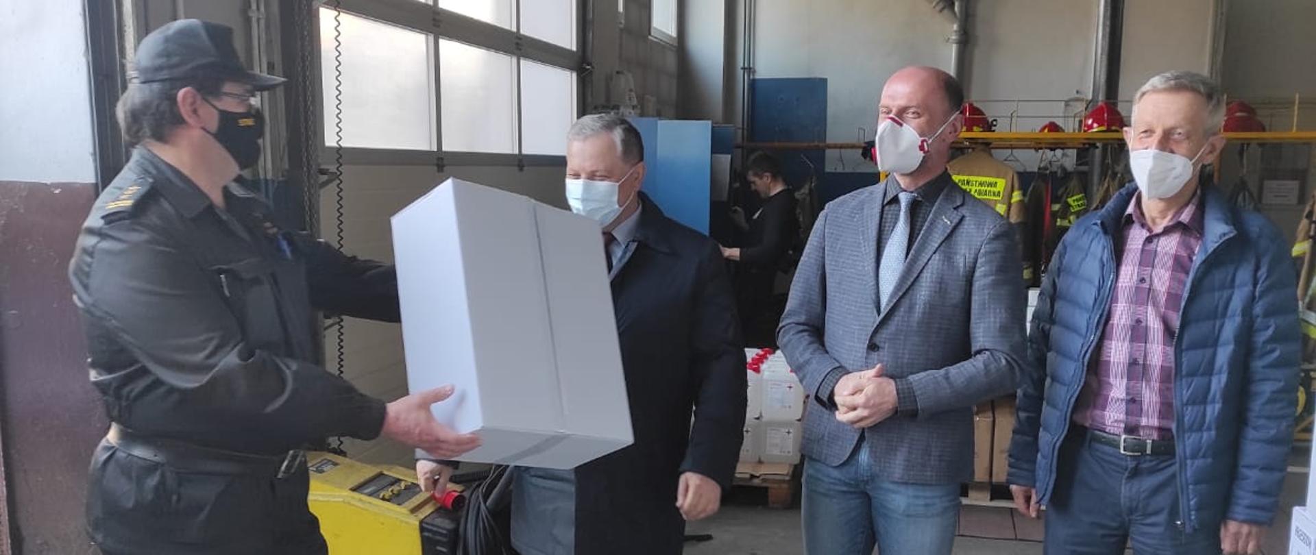 Wnętrze garażu jednostki ratowniczo - gaśniczej. Po lewej stronie Komendant Powiatowy PSP w mundurze sztabowym. Przekazuje karton maseczek przedstawicielom samorz¹du. Po prawej stronie trzech mężczyzn w maseczkach. 