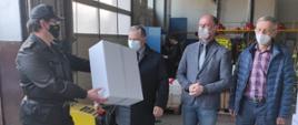 Wnętrze garażu jednostki ratowniczo - gaśniczej. Po lewej stronie Komendant Powiatowy PSP w mundurze sztabowym. Przekazuje karton maseczek przedstawicielom samorz¹du. Po prawej stronie trzech mężczyzn w maseczkach. 