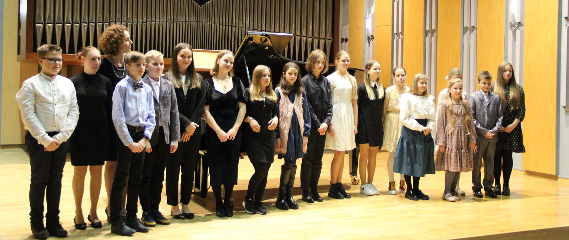 Wykonawcy koncertu "Młodzi pianiści na scenie" Wszyscy wykonawcy wraz z prowadzącą stoją w zwartym szyku na scenie sali koncertowej pozując do grupowego zdjęcia. W tle organy oraz fortepian
