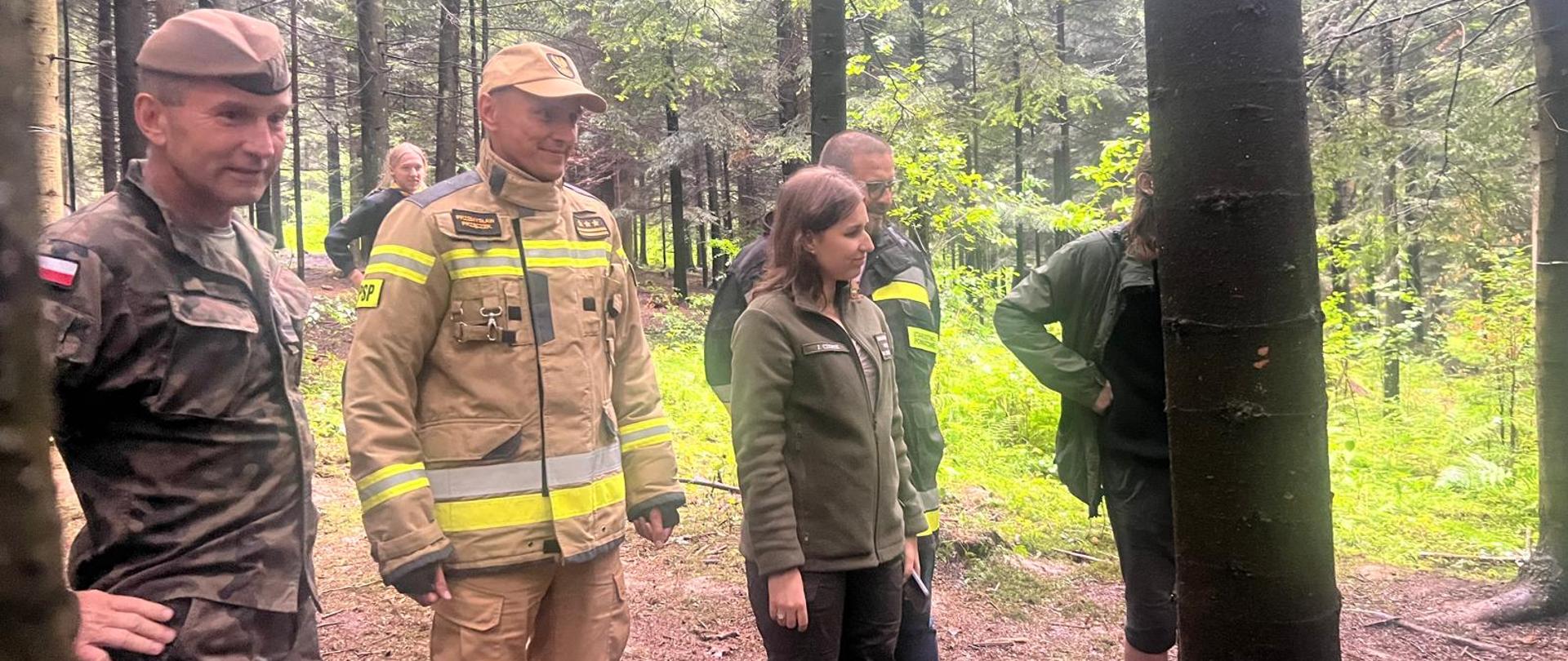 W lesie stoją harcerze w mundurach, oraz komendant wojewódzki w piaskowym ubraniu specjalnym