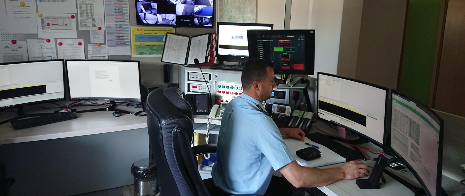 Strażak - Dyżurny Operacyjny Powiatu - za swoim stanowiskiem pracy. Biurko wraz z stajami komputerowymi oraz systemami łączności bezprzewodowej. 