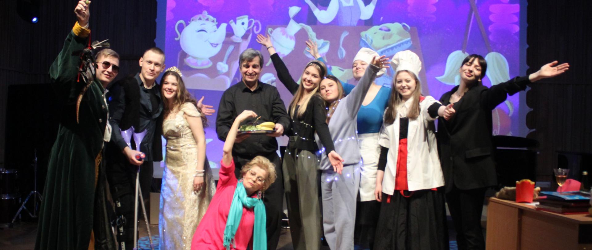 Grafika - zdjęcie. Grupa aktorów po spektaklu na estradzie pozuje do wspólnego zdjęcia. W tle fragment scenografii wyświetlony na ekranie.