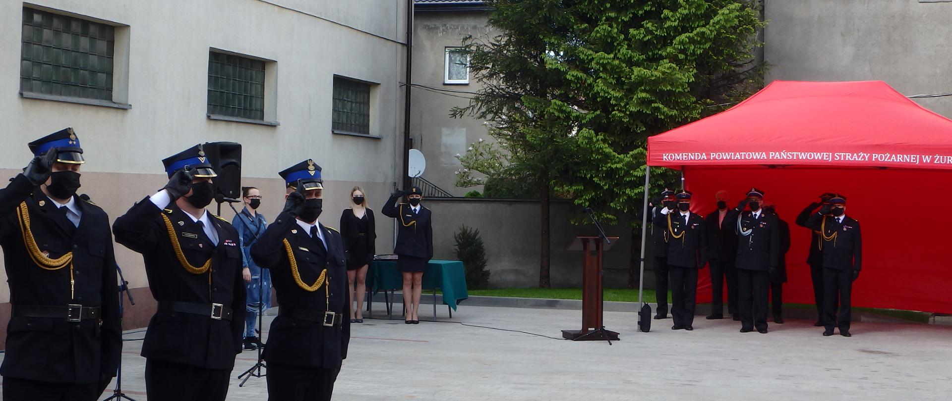 Na placu wewnętrznym komendy odbywają się obchody dnia strażaka. Funkcjonariusze straży pożarnej stoją na baczność salutując.