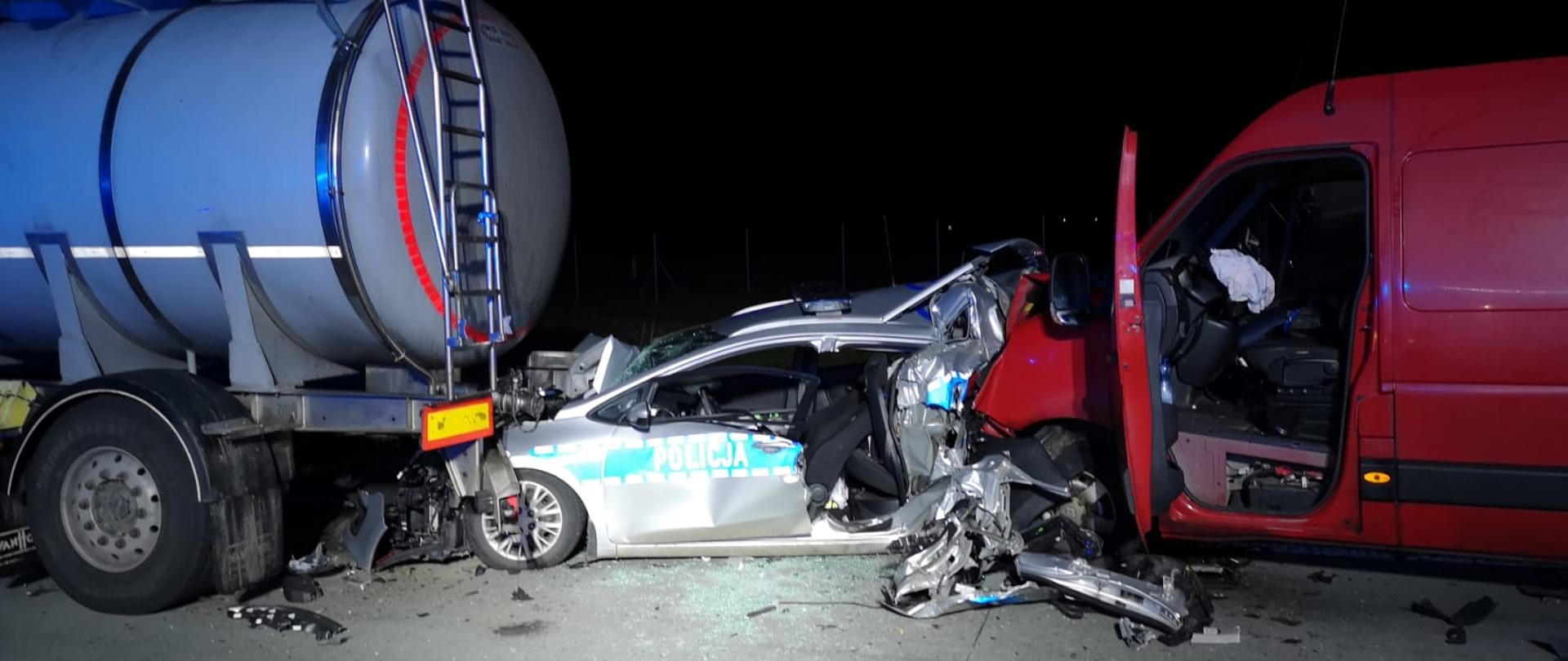 Obraz przedstawia zniszczony radiowóz policyjny zakleszczony pomiędzy busem dostawczym a cysterną. Pojazdy na autostradzie. Pora nocna.