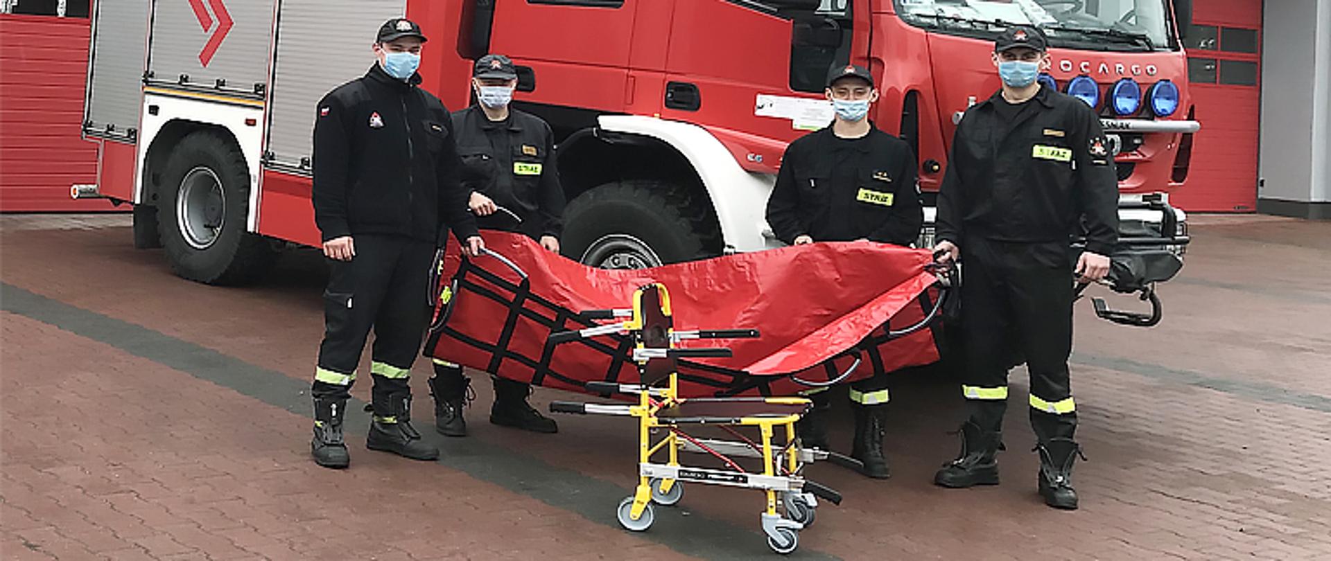 Zdjęcie przedstawia czterech strażaków stojących przed samochodem strażackim trzymających w ręku nosze płachtowe do transportu medycznego koloru czerwonego. Przed nimi stoi specjalistyczne krzesełko do transportu kardiologicznego. Strażacy ubrani są w czarne ubrania koszarowe, na twarzy maja maseczki a na głowie czapkę z daszkiem. W tle widnieje budynek Komendy Powiatowe PSP z dużym napisem Państwowa Straż Pożarna barwy czerwonej, widać bramy garażowe. 