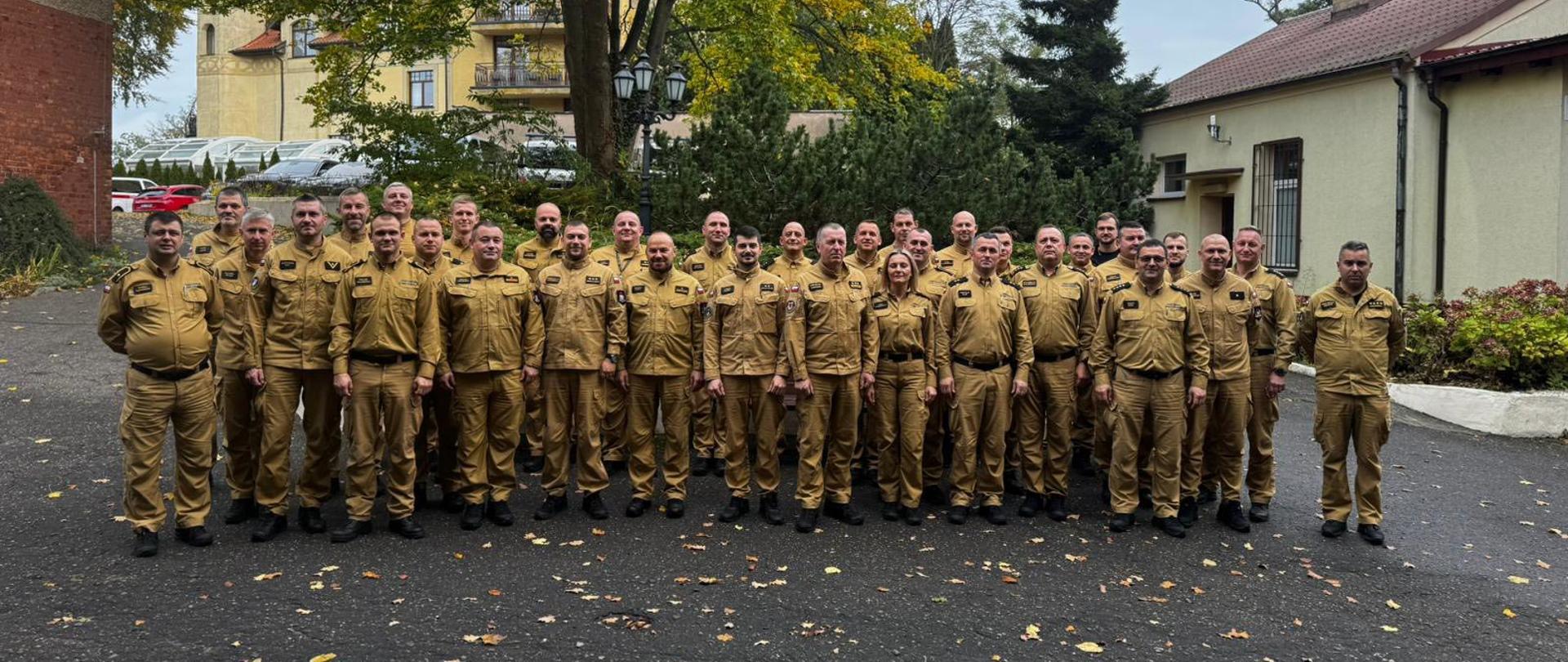 Zdjęcie zrobione na zewnątrz, przedstawiające dużą grupę strażaków, ubranych w identyczne, piaskowe mundury z długimi rękawami i kołnierzami, stojących w kilku rzędach.
Osoby te stoją na brukowanym lub asfaltowym podłożu, usianym opadłymi, jesiennymi liśćmi. W tle widać drzewa z jesiennym listowiem oraz budynki. Wokół panuje pochmurna pogoda.
