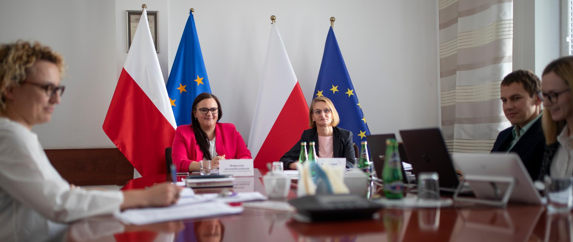Minister Jarosińska-Jedynak siedzi przy stole, za ni ą stoją dwie flagi Polski i dwie UE, przy stole siedzą również pozostali uczestnicy spotkania 