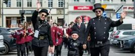 Kobieta, w środku dziecko, następnie mężczyzna - wszyscy w mundurach kominiarskich idą środkiem ulicy. Za nimi orkiestra, w tle kamienice. Po bokach zaparkowane auta osobowe.