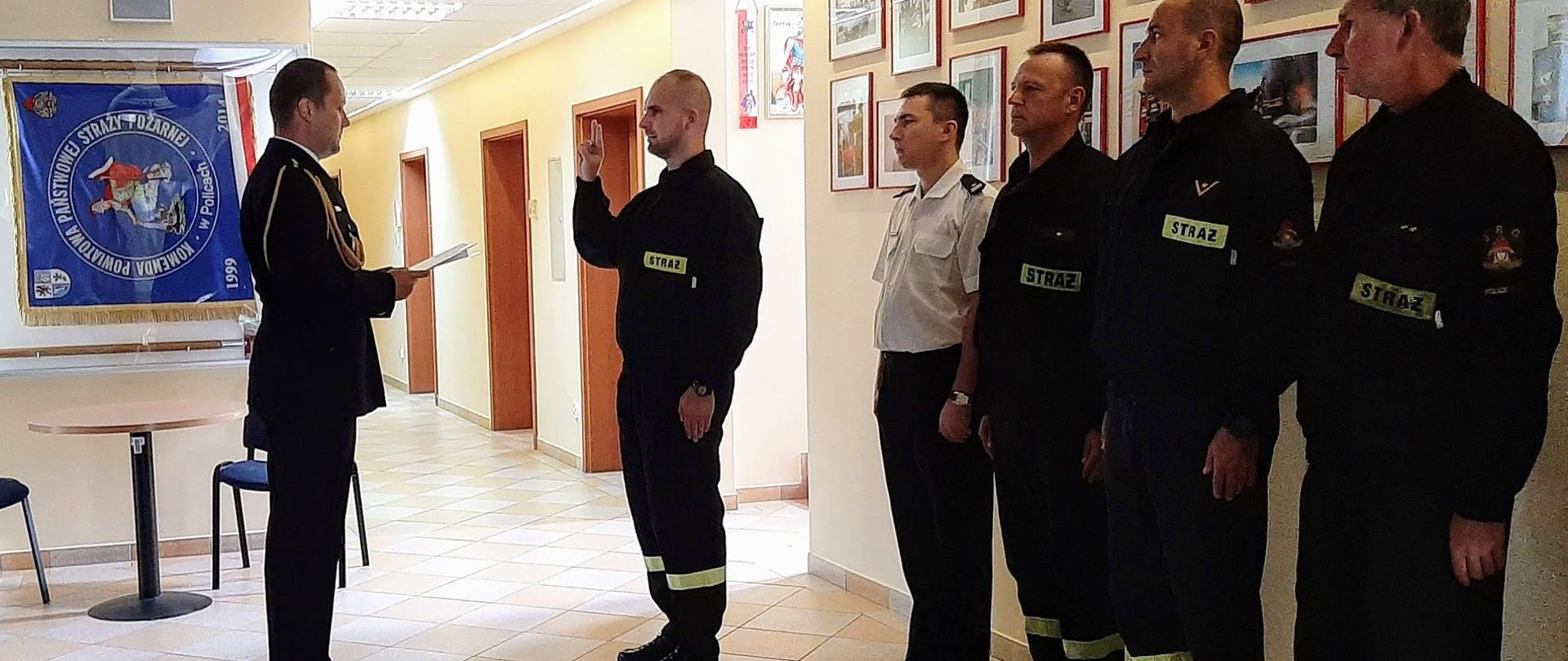 Strażacy stojący na baczność w szergu, jeden stojący przed komendantem, mający podniesioną prawą rękę