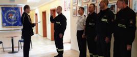 Strażacy stojący na baczność w szergu, jeden stojący przed komendantem, mający podniesioną prawą rękę