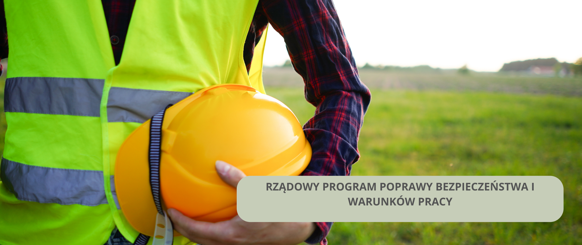 Po lewej stronie widać tors mężczyzny w kamizelce odblaskowej. Mężczyzna trzyma w rękach kask.