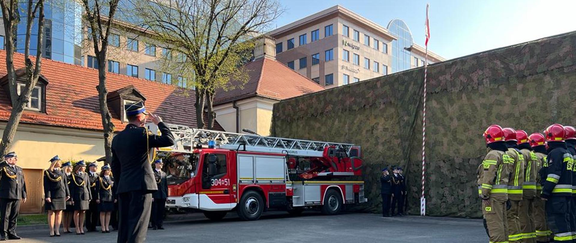 Na zdjęciu widać pododdziały uczestniczące w zbiórce z okazji Dnia Flagi. Na pierwszym planie stoi Dowódca JRG mł. bryg. Dominik Leśniakiewicz w umundurowaniu wyjściowym ze sznurem, który oddaje honor poprzez salutowanie. Z lewej strony zdjęcia widoczny jest pododdział reprezentujący Komendę Miejską z Komendantem Miejskim na czele, który również oddaje honory poprzez salutowanie. Z prawej strony stoją strażacy JRG w umundurowaniu specjalnym. W tle widoczny jest maszt flagowy z wciągniętą do samej góry flagą RP. Na zdjęciu widoczna jest również drabina pożarnicza z numerami operacyjnymi 304 w 51.
