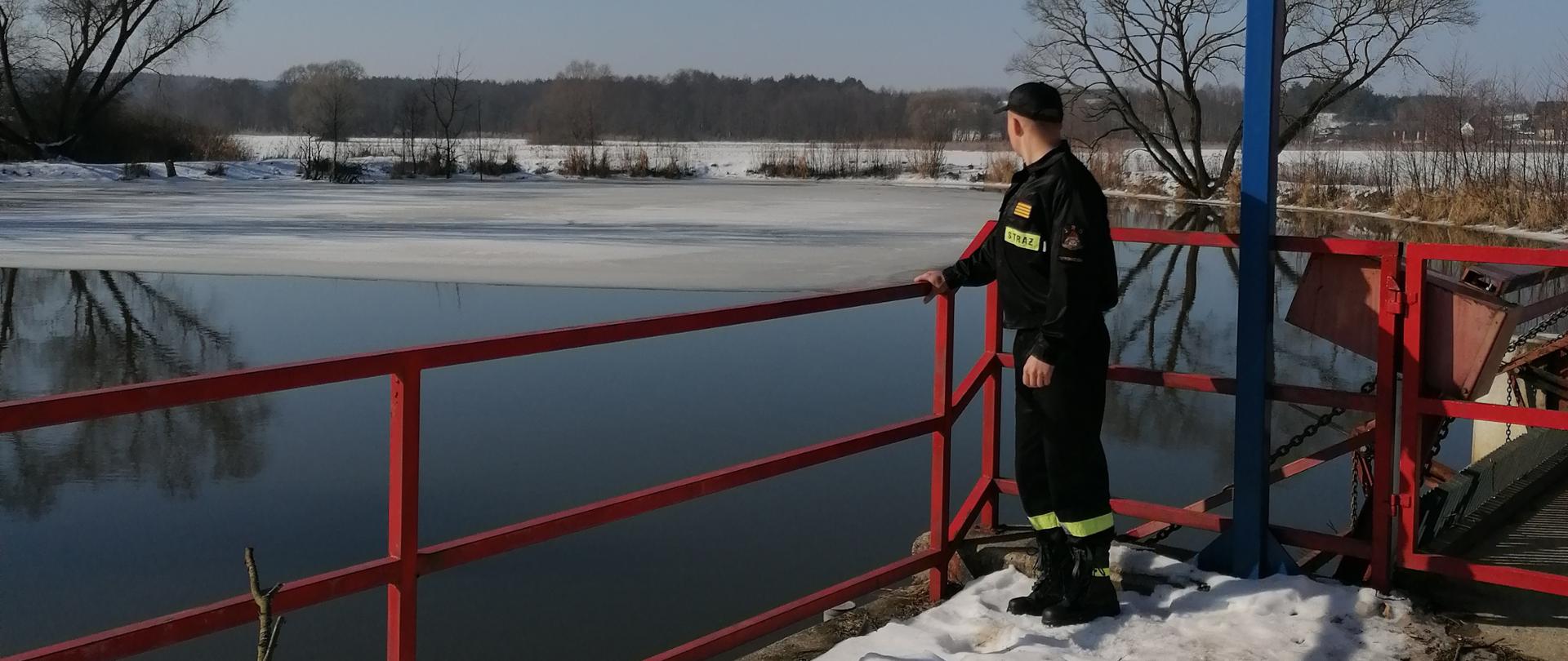 Na pierwszym planie widać stojącego strażaka na pomoście przy rzece Kamienna. Na drugim planie widać zbiornik wodny oraz pobliskie łąki