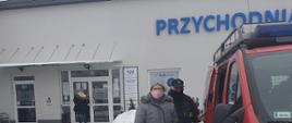 Na zdjęciu widać starszą kobietę w maseczce, z torebką w ręce stojącą razem ze strażakiem ochotnikiem w maseczce obok osobowego samochodu strażackiego zaparkowanego na parkingu przed przychodnią lekarską. Parking częściowo zaśnieżony