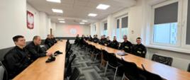 Na zdjęciu widzimy strażaków w czarnych mundurach siedzących w sali wykładowej podczas zajęć teoretycznych. Na ścianie widzimy godło RP Orła Białego w koronie złotej, na drugiej ścianie wyświetlany jest napis Komenda Wojewódzka Państwowej Straży Pożarnej w Kielcach oraz logo PSP.