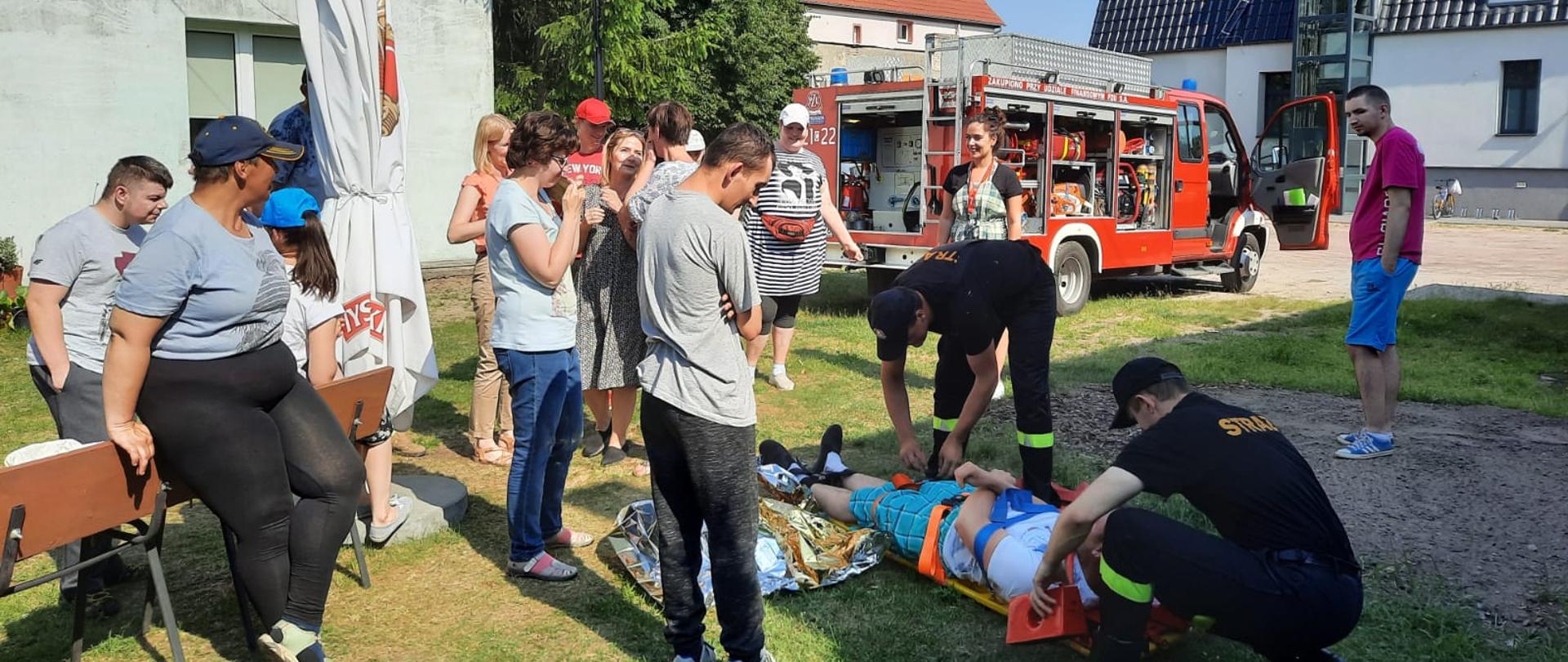 Zdjęcie wykonano podczas pokazów ratownictwa medycznego. Przedstawia na pierwszym planie grupkę osób oraz dwóch strażaków Państwowej Straży Pożarnej z osobą (pozorantem) ułożoną na desce ratowniczej. W tle samochód gaśniczy lekki Państwowej Straży Pożarnej oraz zabudowania.
