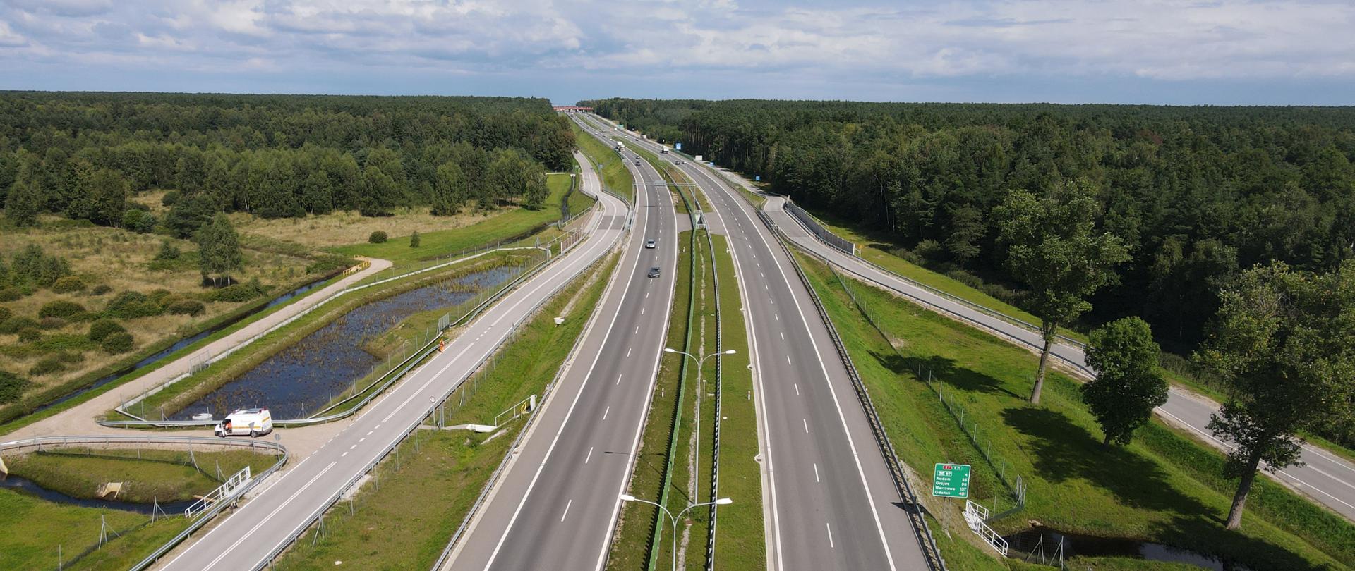 S7 przy granicy województwa świętokrzyskiego i mazowieckiego - dwujezdniowa droga ekspresowa z równoległymi drogami po obu stronach. Po lewej zbiornik retencyjny i ciek wodny. W oddali przejście dla zwierząt pod drogą, a dalej nad drogą. 