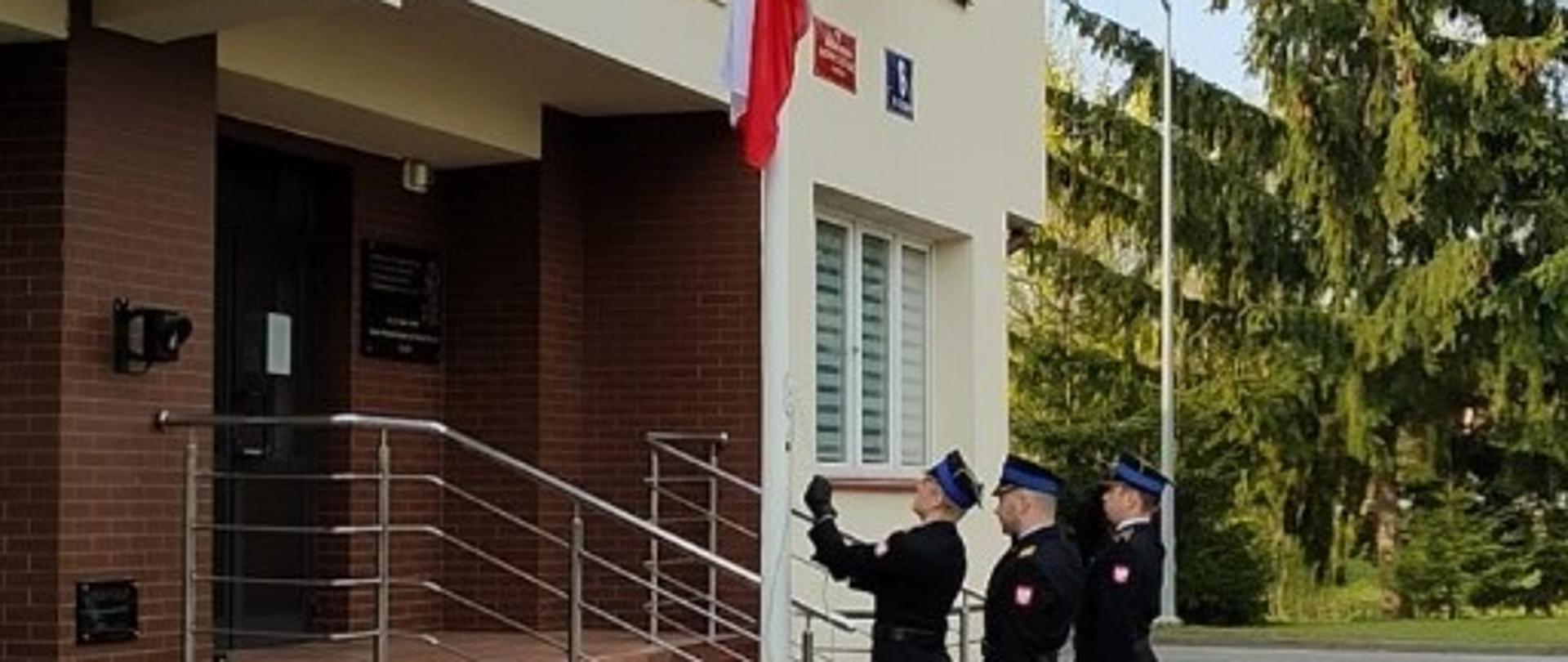 strażacy w umundurowaniu galowy wciągają flagę na maszt. dowódca pocztu i asystujący oddają honor flagowy wciąga flagę