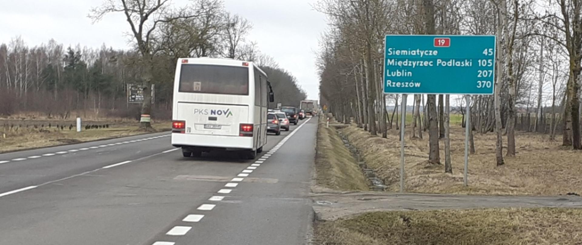 Droga asfaltowa, jedna jezdnia, na jednym pasie ruchu oddalająca się kolumna pojazdów, na pierwszym planie tył autobusu. Obok tablica szlaku drogowego, pokazująca odległość do kolejnych miejscowości 