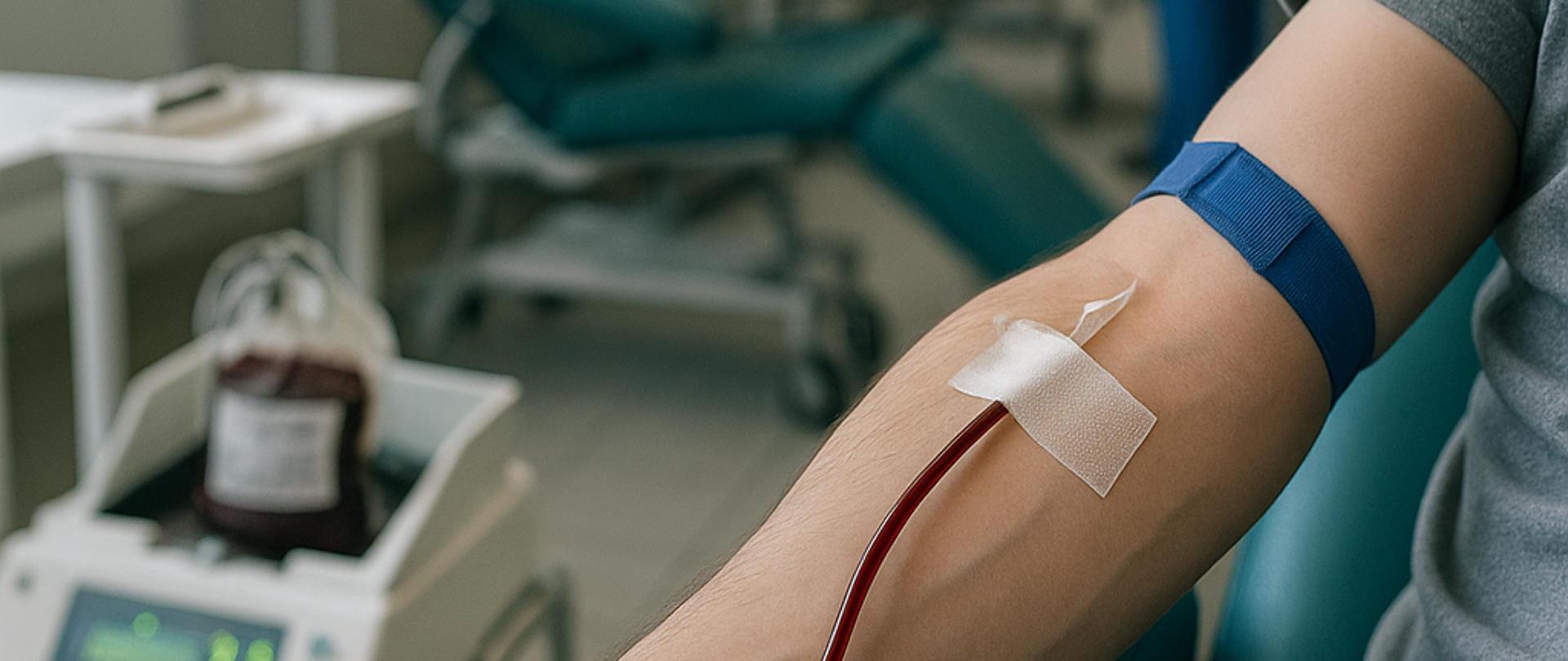 Na zdjęciu widać rękę, od łokcia do końca palców. Do żyły wkłuta jest igła zakończona wężykiem. Jest to moment pobierania krwi. W tle widać pomieszczenie szpitalne gdzie to się odbywa.