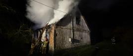 Spalony budynek mieszkalny ze spaloną konstrukcją dachu. Drabina nasadkowa oparta jest o budynek, tworząc stanowisko gaśnicze. Strażacy podają prąd wody do wnętrza budynku. Widoczne zadymienie unoszące się nad budynkiem.