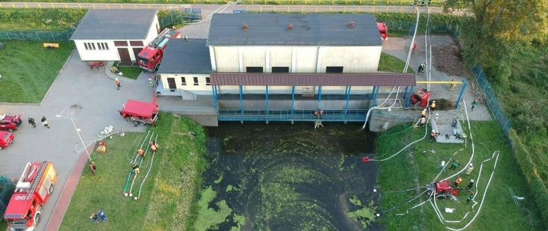 Widok z góry na zbiornik wodny i teren ćwiczeń, obok ustawione samochody gaśnicze PSP, na placu po prawo i lewo i nad zbiornikiem wodnym strażacy przy wężach pożarniczych