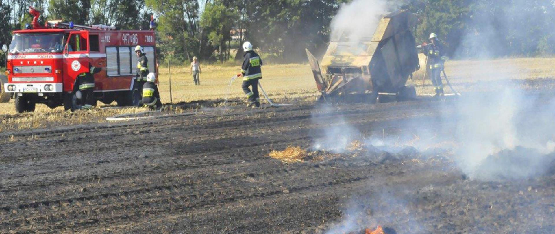 Palące się ściernisko i balot słomy. W tle widać palącą się maszynę rolniczą - balociarkę. Strażacy gaszą pożar. W tle samochód strażacki.