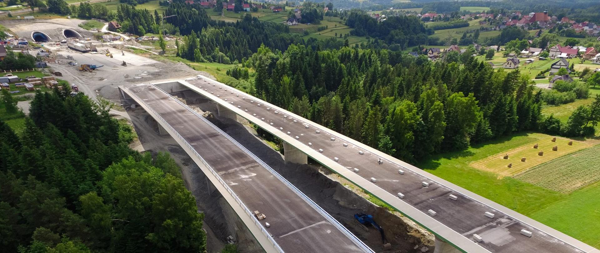 Zdjęcie budowanego mostu przy tunelach. Jezdnie o przeciwnych kierunkach jazdy są oddzielone przestrzenią. Widoczne są maszyny i materiały budowlane na całej długości budowanego mostu, jak również przy tunelach.
Pod mostem przebiega droga serwisowa. Widoczny jest malowniczy krajobraz – dużo wzniesień, dużo zieleni, drzew, piękna panorama.
