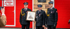 Na tle czerwonej bramy garażowej z napisem ostrzegawczym stoją trzej strażacy w oficjalnych mundurach i czapkach rogatywkach. Dwaj z nich wspólnie trzymają oprawiony w ramę obraz przedstawiający neogotycki kościół. Nad nimi, na filarze budynku, widnieje pamiątkowa tablica poświęcona setnej rocznicy powstania lokalnej jednostki OSP.