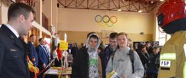 Targi edukacyjne. Sala sportowa w ZSZ w Rawiczu. Przy stoisku Komendy Powiatowej PSP w Rawiczu znajduje się trzech uczniów. Jeden z nich trzyma w ręce folder reklamowy. Z prawej strony widoczny jest fragment manekina mającego na sobie hełm oraz ubranie specjalne. Z lewej strony stoi strażak w mundurze wyjściowym.